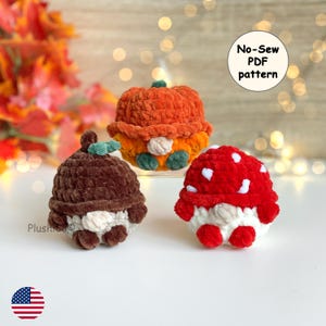 May include: Three small, round, plush crochet figures featuring a brown acorn, an orange pumpkin, and a red mushroom with white spots. Each figure has a soft, textured yarn finish and a small beige nose. A circular label reads No-Sew PDF pattern.