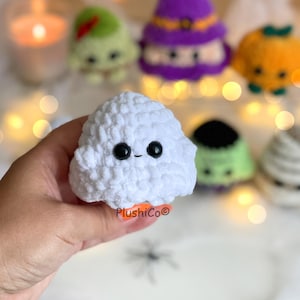 Puede incluir: Un fantasma blanco tejido a ganchillo con ojos de bot&oacute;n negros y una peque&ntilde;a sonrisa. El fantasma es parte de una colecci&oacute;n de peluches de ganchillo con tem&aacute;tica de Halloween, que incluye una bruja, una calabaza y un zombi. El logotipo de Plushico es visible.