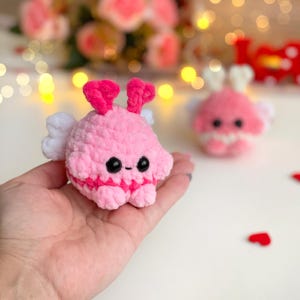 Peut inclure: Une petite peluche ronde en crochet rose clair avec une texture duveteuse. Elle pr&eacute;sente deux antennes en forme de c&oelig;ur rose vif, des ailes blanches sur les c&ocirc;t&eacute;s, ainsi que deux yeux de s&eacute;curit&eacute; noirs et une petite bouche brod&eacute;e.