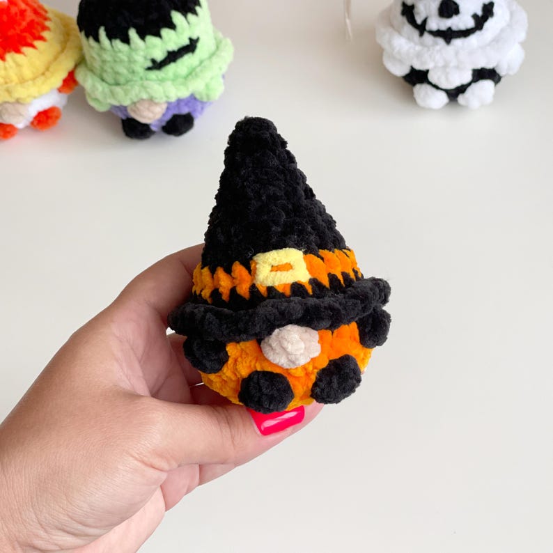 May include: A small, plush, crocheted gnome figure wearing a tall black pointed hat with an orange band and a yellow square buckle. The gnome has a round beige nose, a bright orange body, and small black feet, crafted from soft, fuzzy yarn.