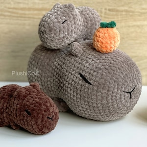 May include: Three crocheted capybara plushies. Two are gray and one is brown. The gray capybaras are stacked on top of each other. The brown capybara is lying on its side. A small orange crocheted fruit is on top of the gray capybaras.