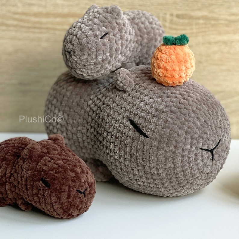 May include: Three crocheted capybara plushies. Two are gray and one is brown. The gray capybaras are stacked on top of each other. The brown capybara is lying on its side. A small orange crocheted fruit is on top of the gray capybaras.