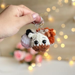 May include: A handmade crochet keychain featuring a small, plush cow with a white face, brown muzzle, and black and white ears. The cow has large, black eyes and is attached to a silver keyring. The keychain is a cute accessory.