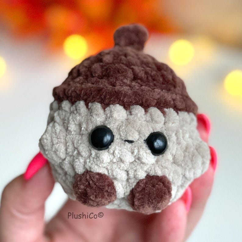 May include: A handmade, plush acorn toy with a light gray body and a dark brown cap. The acorn has two large, black, glossy eyes and small brown feet. The toy is held in a hand with pink nail polish. The brand name "PlushiCo" is visible.