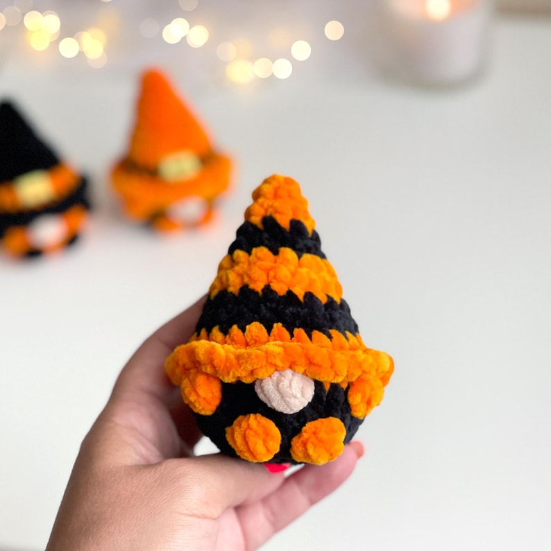 May include: A small, plush, crocheted gnome figure featuring a pointed hat with alternating horizontal stripes of black and orange yarn. The gnome has a round, light-colored nose and a black body with orange accents, crafted from soft, fuzzy material.