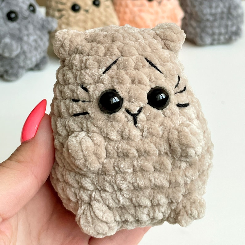 May include: A plush beige cat toy with black embroidered eyes and whiskers. The cat toy is made of a soft, fuzzy material.