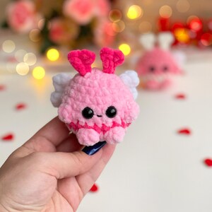 Peut inclure: Un petit jouet en peluche crochet&eacute;, rond et rose, avec une texture douce et duveteuse. Il pr&eacute;sente deux antennes en forme de c&oelig;ur rose vif sur le dessus, des ailes blanches sur les c&ocirc;t&eacute;s et deux yeux de s&eacute;curit&eacute; noirs en plastique.