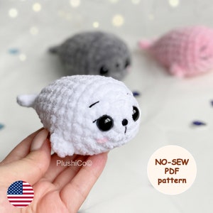 May include: A white crocheted seal plushie with large black eyes and a small pink blush. The image shows three plush seals, with a gray and pink seal in the background. The text "NO-SEW PDF pattern" is in the image.