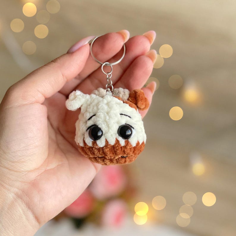 May include: A handmade crochet keychain in the shape of a dog's head. The keychain features a white and brown color scheme, with large black eyes. The dog's head is attached to a silver keyring.