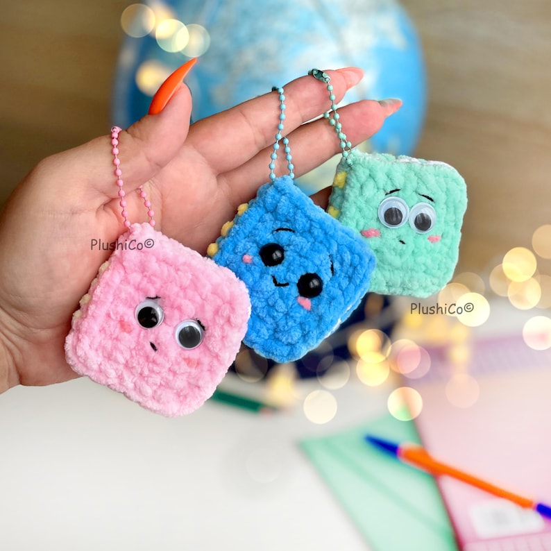 May include: Three plush, square-shaped keychains in pink, blue, and green, each with googly eyes and a stitched mouth. The keychains have a chain and the text "PlushiCo" is visible. The blue keychain has yellow trim.