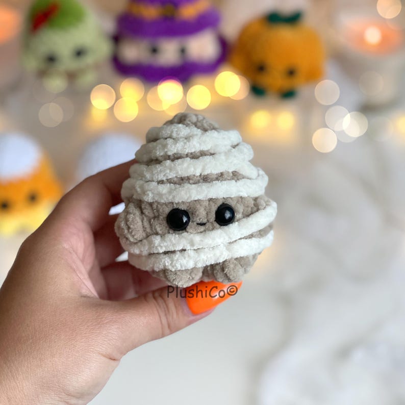 Puede incluir: Un juguete de peluche con forma de momia, de color marr&oacute;n claro, con una cara sonriente y envuelto en hilo blanco. La momia tiene ojos negros de bot&oacute;n. Otros juguetes de peluche con tem&aacute;tica de Halloween son visibles en el fondo.