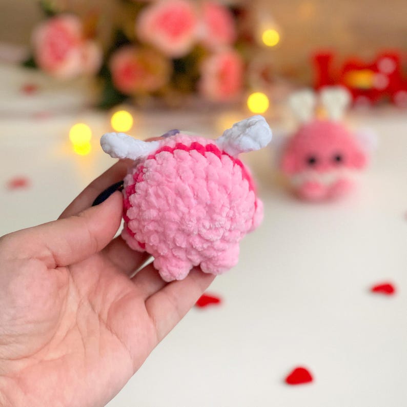Peut inclure: Un petit jouet en peluche crochet&eacute; rose et rond, dot&eacute; d'ailes en fil blanc doux fix&eacute;es sur le dessus. Le fil textur&eacute; et duveteux forme une silhouette arrondie avec une bande horizontale distincte de fil rose fonc&eacute; sur le haut.