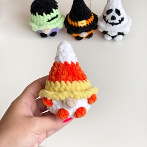 May include: Four small, plush, crocheted figures featuring soft yarn textures. One figure is shaped like candy corn with white, orange, and yellow stripes. Three other figures in the background include a green monster, a black witch, and a white ghost.