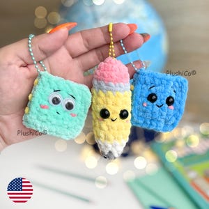 May include: Three handmade crochet keychains: a teal square, a yellow pencil, and a blue square, each with googly eyes and smiling faces. The pencil has a pink eraser and a gray tip. Each keychain is attached to a metal chain. The brand name "PlushiCo" is visible.