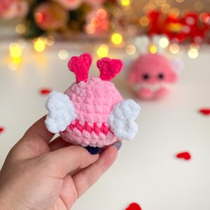 Peut inclure: Un petit jouet en peluche crochet&eacute;, rond et rose clair, dot&eacute; de deux antennes en forme de c&oelig;ur rose vif sur le dessus et de deux ailes en forme de c&oelig;ur blanc sur les c&ocirc;t&eacute;s. La peluche a une finition en fil textur&eacute; et une bande horizontale rose fonc&eacute;.