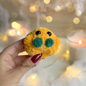 May include: A handmade, plush toy in a vibrant yellow color, featuring two green circular accents and black eyes. The plushie has a textured, soft appearance and is held in a hand. The brand name "PlushiCo" is visible.