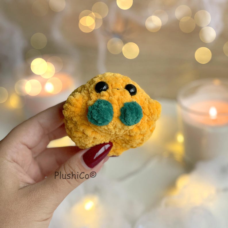 May include: A handmade, plush toy in a vibrant yellow color, featuring two green circular accents and black eyes. The plushie has a textured, soft appearance and is held in a hand. The brand name "PlushiCo" is visible.