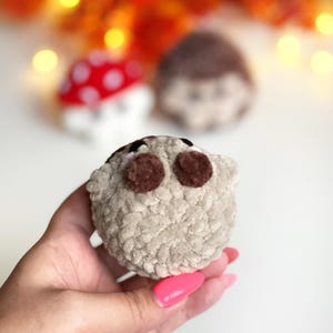 May include: A handmade, plush, light gray hedgehog toy with brown eyes and a small black nose. The toy is held in a hand with pink nail polish. Two other plush toys are in the background, one a mushroom with red and white spots.