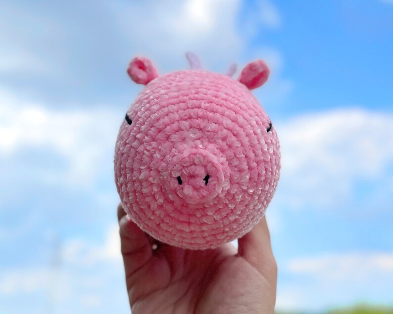 K&ouml;nnte beinhalten: Ein rosa geh&auml;keltes Schweinchenspielzeug mit schwarzen Augen und einer kleinen Schnauze. Das Spielzeug wird in einer Hand vor einem blauen Himmel gehalten.