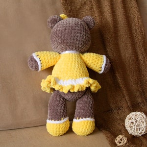 May include: A brown crocheted teddy bear wearing a yellow crocheted dress with white trim. The bear is sitting on a brown fabric surface.