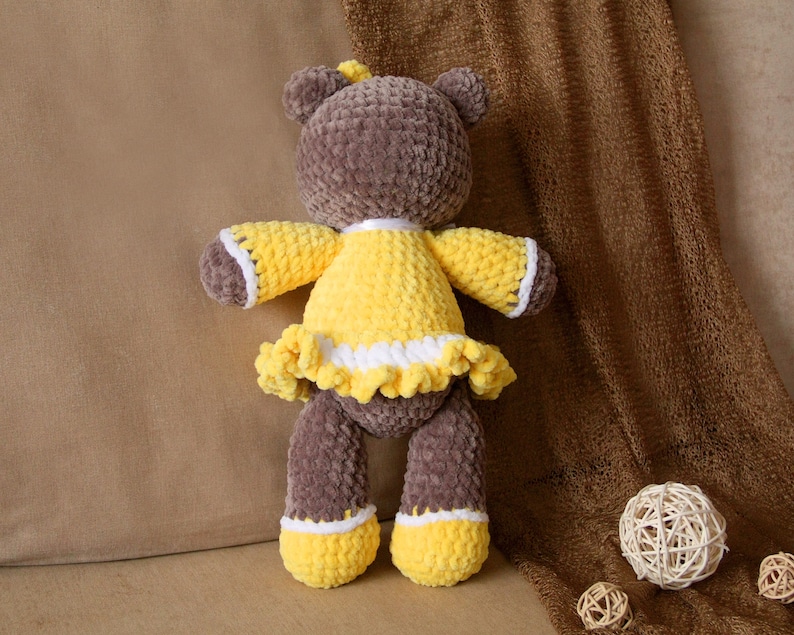 May include: A brown crocheted teddy bear wearing a yellow crocheted dress with white trim. The bear is sitting on a brown fabric surface.