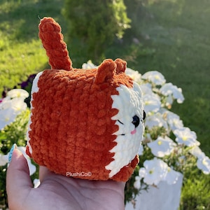 May include: A crocheted plush toy shaped like a fox with orange fur and white facial features. The toy is being held in a hand.