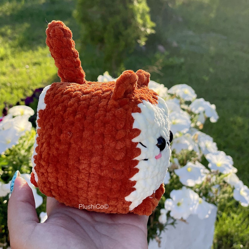 May include: A crocheted plush toy shaped like a fox with orange fur and white facial features. The toy is being held in a hand.