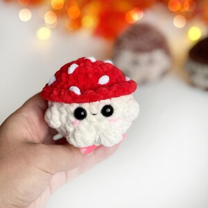 May include: A handmade crochet mushroom toy with a red cap dotted with white spots. The mushroom has a white body, black eyes, and pink cheeks. The toy is held in a hand, with other similar toys blurred in the background.