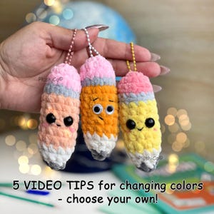 May include: Three plush pencil keychains in peach, orange, and yellow, each with a pink eraser, gray tip, and black eyes. The keychains are held up by a chain. The text on the image says "5 VIDEO TIPS for changing colors - choose your own!"