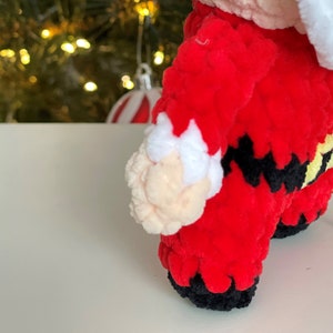 May include: A crocheted Santa Claus figure made of soft red, white, black, and yellow yarn. The figure features a red suit with white trim, a black belt with a yellow buckle, and black boots. The texture is plush and fuzzy throughout the piece.