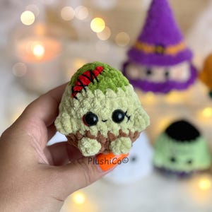 Puede incluir: Un cupcake zombi de peluche hecho a mano, con una cobertura verde, puntadas rojas y ojos negros. El cupcake se sostiene en una mano, con otros peluches tem&aacute;ticos de Halloween en el fondo. El cupcake tiene una base marr&oacute;n y una boca cosida.