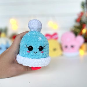 May include: A small, light blue crocheted plush toy shaped like a winter hat with a white pom-pom on top and a white brim. The hat features embroidered black eyes, long eyelashes, a small black mouth, and pink cheeks, with white star accents.