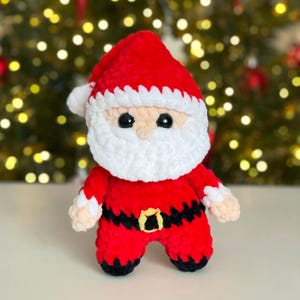 May include: A handmade crochet Santa Claus doll crafted from soft red, white, and black yarn. The figure features a plush red hat with a white brim, a thick white beard, black safety eyes, and a black belt with a yellow square buckle.