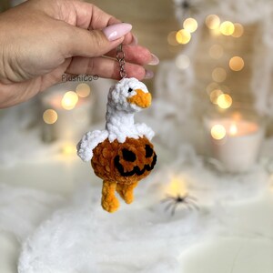 May include: A handmade crochet keychain featuring a goose with a pumpkin body. The goose has a white head and neck, an orange beak, and a pumpkin-shaped body with a carved face. The keychain is held by a silver keyring.