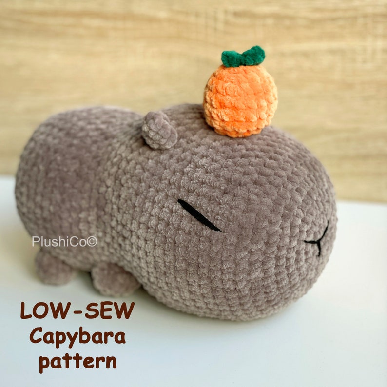 May include: A crocheted gray capybara plush toy with a small orange crocheted fruit on its head. The toy is lying on a white surface. The text "LOW-SEW Capybara pattern" is below the toy.