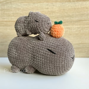 May include: Two gray crocheted capybara plushies, one on top of the other, with an orange crocheted fruit on top of the smaller capybara.
