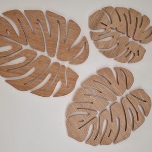 Può includere: Tre pezzi di arredamento da parete in legno a forma di foglie di monstera. Le foglie sono di dimensioni diverse e sono realizzate in legno marrone chiaro.
