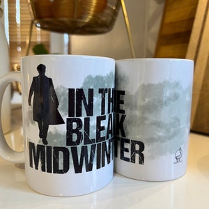 May include: Two white ceramic mugs with black text and a silhouette of a person. The mug on the left says "In the Bleak Midwinter". The mug on the right has a watercolor-like design of clouds.