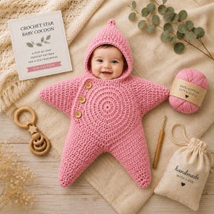 May include: A pink crocheted star-shaped baby cocoon with a hood and three wooden buttons. The set includes a ball of pink cotton yarn, a wooden crochet hook, a wooden rattle, a small drawstring bag labeled handmade with love, and a pattern card.