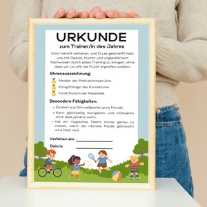 Może przedstawiać: Certyfikat z niebieskim tłem i drewnianą ramą. Certyfikat nosi tytuł "URKUNDE zum Trainer/in des Jahres" i zawiera listę nagród i umiejętności. Certyfikat jest ilustrowany dziećmi bawiącymi się na zewnątrz.