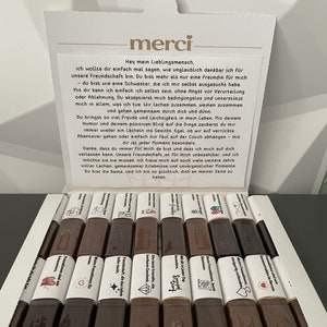Puede incluir: Una caja blanca con un logotipo merci rojo y blanco. La caja contiene una variedad de barras de chocolate con diferentes mensajes impresos en ellas. Los mensajes están en alemán e incluyen frases como "Ein herz für Freunde" y "Sonnenstrahlen im Herzen".