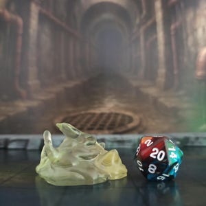 Puede incluir: Una figura de criatura amorfa, translúcida y de color amarillo pálido, con una forma suave y orgánica, colocada junto a un dado poliédrico de veinte caras multicolor con números blancos, incluido un 20 prominente en su cara superior.