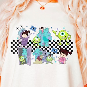 May include: White t-shirt with a checkered pattern and a graphic of characters from the movie Monsters Inc. The characters include Sully, Mike, Boo, and Randall. The graphic is in a cartoon style and features bright colors.