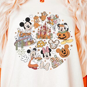 Puede incluir: Una camiseta blanca con un estampado gráfico circular de varios personajes e iconos de Disney, incluyendo a Mickey Mouse, Minnie Mouse, Epcot, un castillo, calabazas y el texto "It's a small world" en una paleta de colores otoñales.