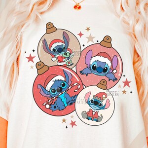 May include: White t-shirt with a Christmas-themed design featuring four Stitch characters from the Disney movie Lilo & Stitch. The characters are inside of red, pink, and beige ornaments with gold glitter accents. The text "bloom pattern" is visible in the bottom left corner of the image.