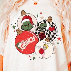 以下が含まれることがあります： クリーム色のTシャツ。クリスマスオーナメントのデザインで、GrinchとCindy Lou Whoが描かれています。Grinchのオーナメントはサンタ帽と白黒の市松模様です。「The Grinch」の文字が赤いオーナメントに書かれています。