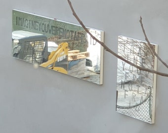 May include: Two rectangular mirrors mounted on a white wall. The mirror on the left reflects a yellow construction vehicle and the text "IMAGINE YOU WERE NOT A" in black. The mirror on the right reflects a blurry image of a fence and the text "YOU WERE NOT A" in black.