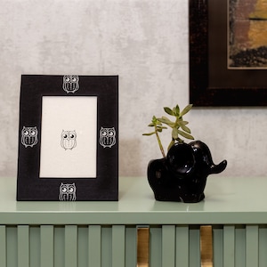 Pode incluir: Moldura de foto preta com um design de coruja branca na borda. A moldura está sobre uma prateleira verde claro com um vaso de elefante preto com uma planta suculenta verde.