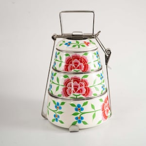 Op de afbeelding: Een drielaagse, witte metalen lunchbox met een bloemenontwerp. De lunchbox heeft een rood en roze pioenroosontwerp, met blauwe en groene accenten. Een metalen handvat en sluitingen zekeren de gestapelde containers.