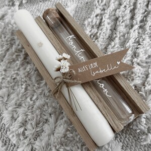 Puede incluir: Una vela blanca y un vial de vidrio con la palabra "Konfir" se muestran en un soporte de madera. Se adjunta una peque&ntilde;a etiqueta con las palabras "Alles Liebe Isabella". Decorado con flores secas.